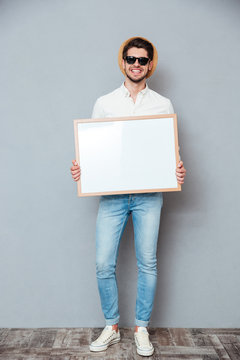 Smiling young man in hat and sunglasses holding white board