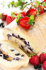 Biscuit roll with mascarpone cream and blueberries decorated strawberries, blueberries and mint leaves on white wooden background