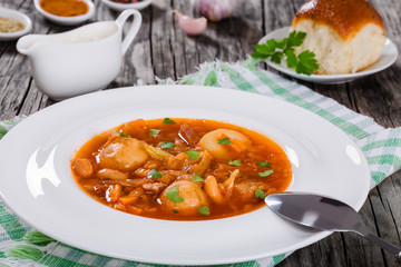 Traditional Ukrainian or Russian vegetable soup, borscht cooked in meat broth with new potatoes  in white wide rim dish. sour cream in gravy boat and bun sprinkled with garlic on background, close-up