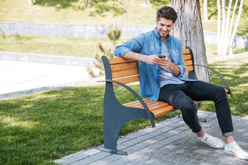 Happy man sitting on the bench outdoors and using smartphone