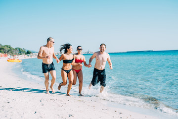 Group of multiethnic man and woman friends in summertime running on the beach hand in hand, smiling - friendship, summer, joyful concept