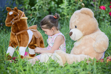 Little girl playing with toys, Teddy bear and horse, sitting on green grass outside in backyard in a sunny summer day