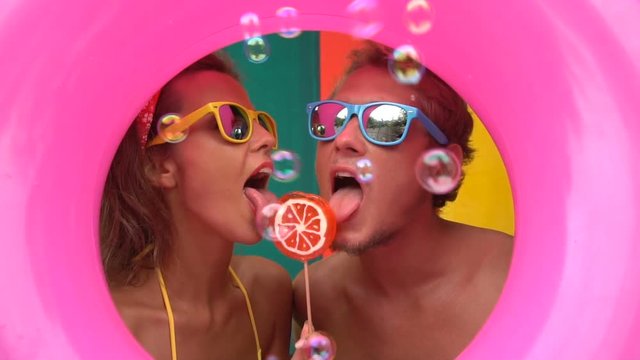 Funny young couple eating soap bubbles and licking orange lollipop isolated in the middle of pink inflatable ring and colorful wall in slow motion.