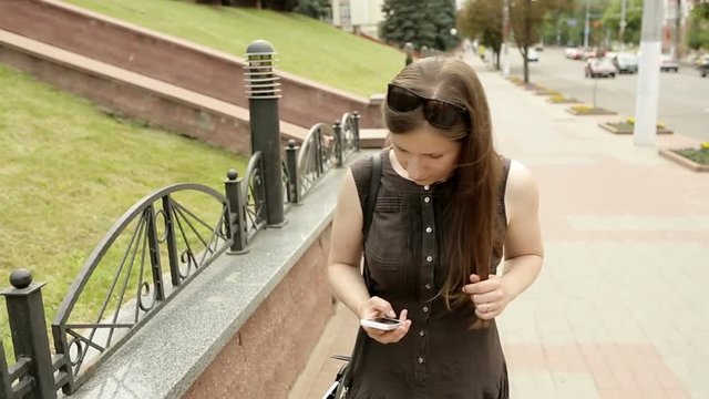 Girl in brown dress walking and texting on the smart phone in the street at sunny weather. Steadycam shoot