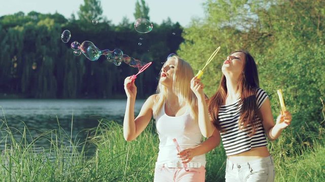 Funny girls blowing colourful soap bubbles and rejoicing near the lake. Slowly