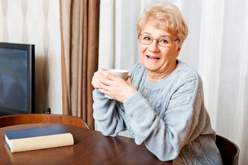 Senior woman sitting at the desk with book and drinking tea or coffee