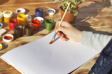 The artist's hand holds the brush and empty album, paint, pencils and flower on wooden background. The view from the top