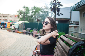 Girl in sunglasses sitting on a wooden bench in the city and waiting for friends. One girl waiting for friends.