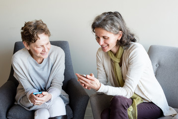 Older mother and middle aged daughter seated next to each other using smart phones and smiling