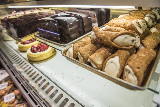 Cannoli and slices of cakes on dessert display behind glass in a store