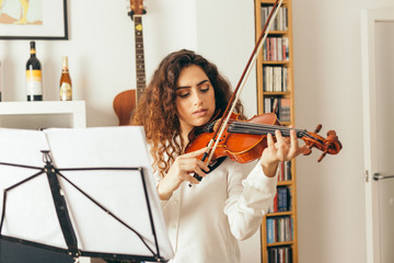 Girl playing violin. Young woman studying music alone at home in the living room with natural and soft light. Curly long and brunette hair, elegant dressed.