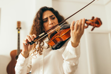 Girl playing violin. Young woman studying music alone at home in the living room with natural and soft light. Curly long and brunette hair, elegant dressed.
