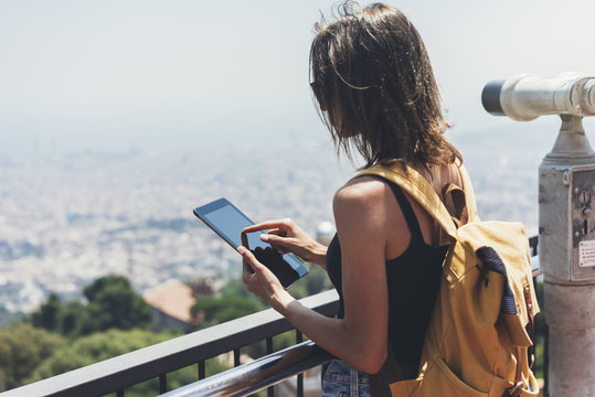 Hipster young girl with bright backpack planning travel plan. Tourist traveler on background panoramic view city, binoculars. Mock up for text message. Female hands using tablet, holding gadget