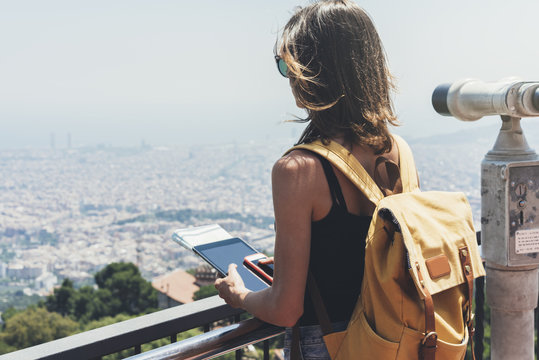 Hipster young girl with bright backpack. Tourist traveler on background panoramic view of the city, binoculars. Mock up for text message. Barcelona Tibidabo. Female hands using tablet gadget