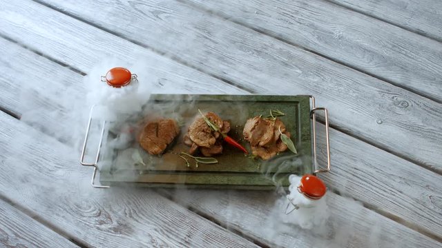 Pieces of meat on tray. Woman's hand puts small jar. Veal medallions with herbs. Food served with dry ice.