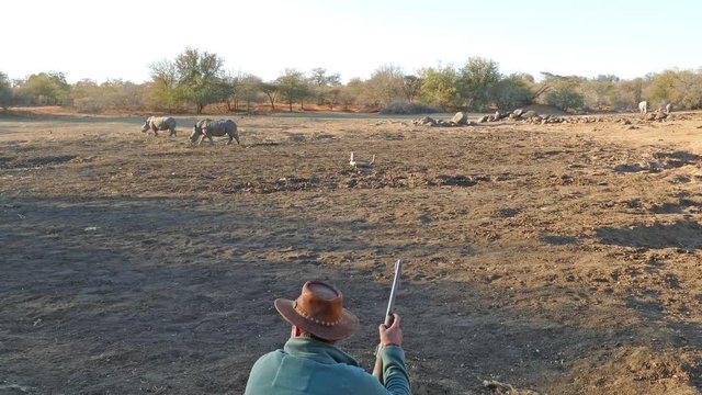 Ranger with firearm observes rhinos at dry water hole.
