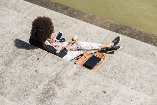 Young woman sitting on stairs eating an apple while looking at her smartphone