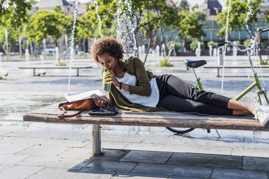 Young woman lying on a bench drinking beverage while looking at her smartphone