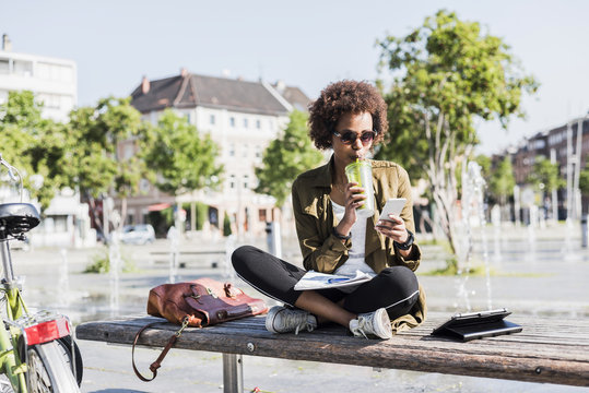 Young woman sitting on a bench drinking beverage while looking at her smartphone