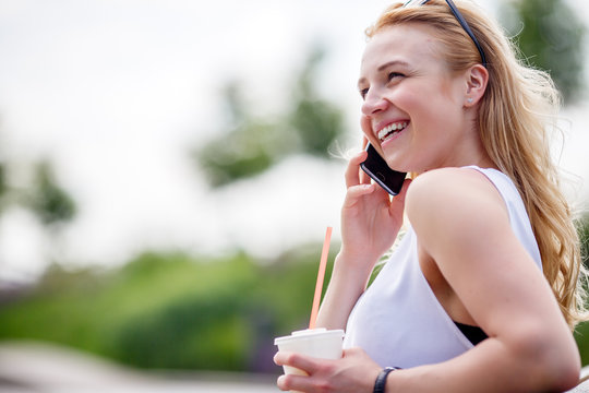 Blonde girl with coffee talking on phone in park bench