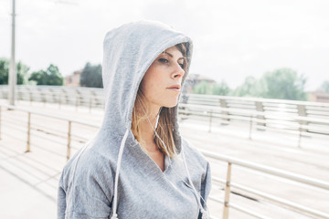 Portrait of a young woman while resting after physical workout outdoors. She wears hooded...
