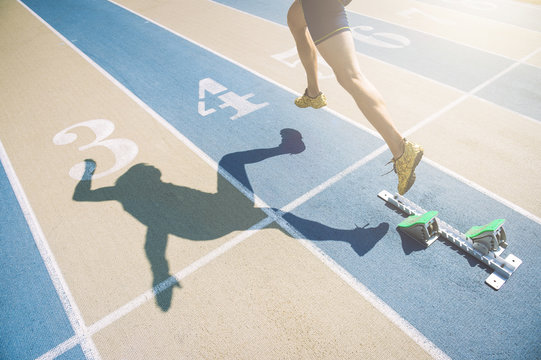 Athlete in gold shoes sprinting from the blocks over the starting line of a race on a blue and tan running track 