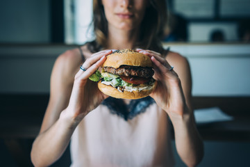 Young girl holding in female hands fast food burger, american unhealthy calories meal on blue background, mockup with space for text message or design, hungry human with grilled hamburger front view