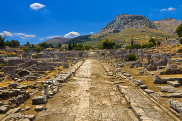 Greece. The Archaeological Site of Ancient Corinth. Lechaion Road (cardo maximus) paved with limestone slabs and remains of monuments. There is Acrocorinth with fortified citadel in the background