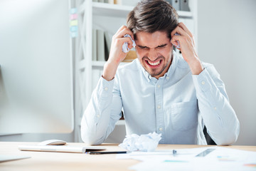 Aggressive angry businessman holding crumpled paper sitting at workplace