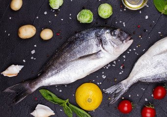 Raw fresh dorado fish with brussels sprouts, tomatoes, lemon, young potato, greens, bread, white wine bottle and olive oil on dark background. View from above, top studio shot