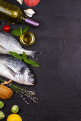 Raw fresh dorado fish with brussels sprouts, tomatoes, lemon, young potato, greens, bread, white wine bottle and olive oil on dark background. View from above, top studio shot