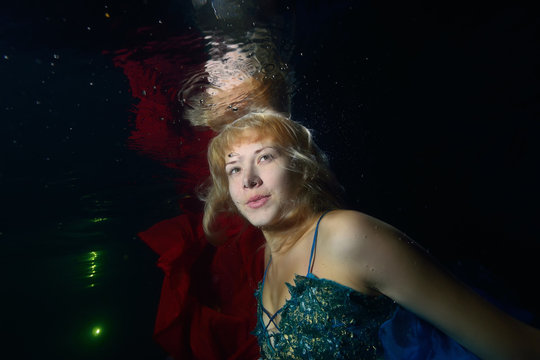 A girl is swimming and posing underwater in a blue dress with red fabric on a black background. Scruffy hair. Close-up. Portrait. Horizontal orientation.