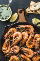 Roasted tiger prawns in iron pan on wooden board with fresh leek, lemon slices, bread and pesto sauce over black background