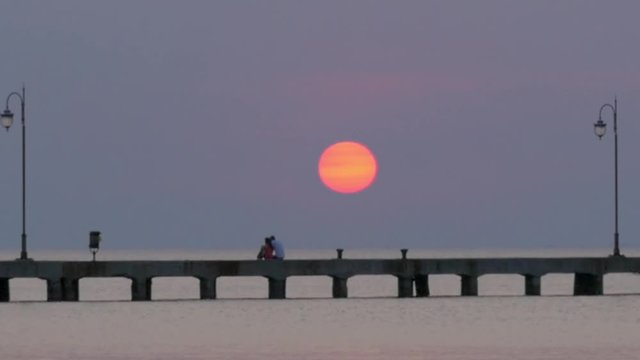 Cinemagraph - Orange sun is setting in clouds over the sea, couple is resting on the pier