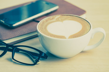 A cup of coffe with heart pattern in a white cup on, smartphone, leather notebook and eyeglasses on wooden  table.Close up.Vintage tone