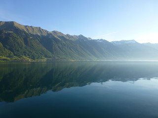 View across Brienzersee Lake, Switzerland on a sunny morning in late Spring, with the surrounding mountains reflected in the calm waters of the lake