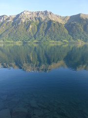 Fototapeta na wymiar Early morning shot of Brienzersee lake, Switzerland, with mountain range opposite reflected in the calm lake, and submerged rocks visible through the clear water in the foreground