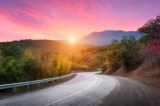 Fototapeta Mountain road passing through the forest with dramatic colorful sky and red clouds at colorful sunset in summer. Mountain landscape with road. travel background