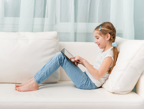 little girl sitting on white sofa looks at tablet touching it profile photo Stock Adobe Stock