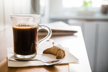 Black coffee in glass with spoon and croissant on wooden table