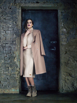 Elegant fashion model wearing long mocha coat, posing before a door.