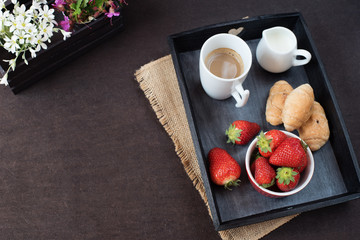 Coffee, mini French pastries and strawberries on wooden tray over black table. White and purple flowers in a decorative wooden crate. Black background