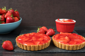 Chocolate tart, tartalette with white chocolate and mascarpone cream, fresh strawberries on top. Large blue bowl full of strawberries. Black wooden background.