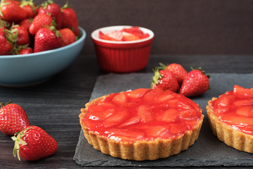 Chocolate tart, tartalette with white chocolate and mascarpone cream, fresh strawberries on top. Large blue bowl full of strawberries. Black wooden background.