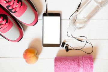 Red sneakers with towel, peach, bottle with water, smartphone with copy space on screen with headphones on white wooden background indoors. Mobile phone mockup.
