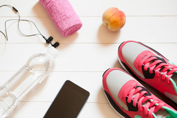 Red sneakers with towel, peach, bottle with water, smartphone with headphones on white wooden background indoors. Sports and fitness background, healthy lifestyle concept.