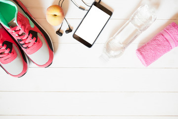 Red sneakers with towel, peach, bottle with water, smartphone with copy space on screen with headphones on white wooden background indoors. Mobile phone mockup.