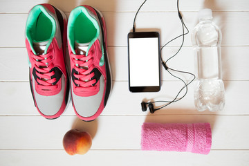 Red sneakers with towel, peach, bottle with water, smartphone with copy space on screen with headphones on white wooden background indoors.