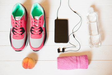 Red sneakers with towel, peach, bottle with water, smartphone with headphones on white wooden background indoors. Sports and fitness background, healthy lifestyle concept.