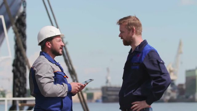 Two skilled workers at a port with a tablet PC, nearby cargo ship partly visible